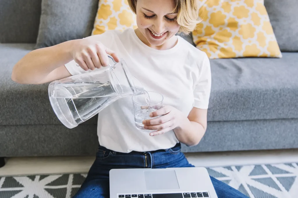 A smiling woman with a laptop pouring water into a glass, illustrating the importance of staying hydrated to manage diabetes and prevent teeth problems.