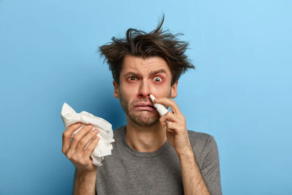 Young man suffering from allergies with nasal spray and tissues, illustrating the impact of allergies on oral health